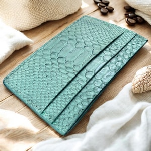 Puede incluir: Un tarjetero de color turquesa con un patrón de piel de serpiente. La cartera de cuero tiene tres ranuras para tarjetas y se exhibe sobre una superficie de madera. El fondo incluye tela blanca, un sombrero tejido y granos de café.