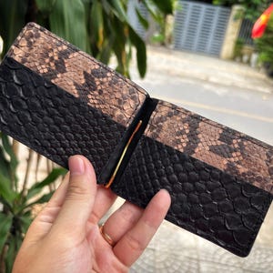 May include: Open leather wallet with a bi-color design. The bottom half is black, and the top half is brown with a snakeskin pattern. A gold-colored money clip is visible inside the wallet.