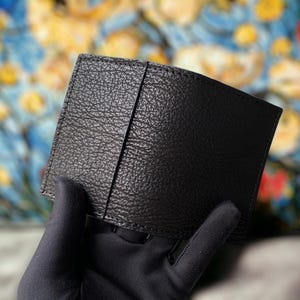 May include: A black leather wallet with a textured finish. The wallet is held by a gloved hand. The wallet appears to be new. The background is a colourful, blurred image.