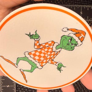 May include: A circular sticker featuring a cartoon Grinch character in a Santa hat and checkered orange and white outfit. The Grinch is green with a mischievous expression, set against a white background with an orange border.
