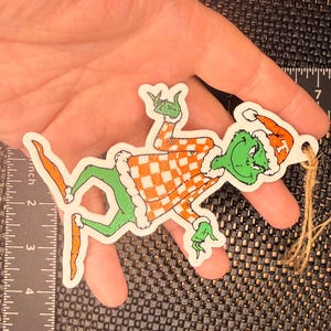 May include: A wooden Christmas ornament featuring a cartoon Grinch character. The Grinch is green with a Santa hat and an orange and white checkered outfit. The ornament is approximately 4 inches long and has a jute string for hanging.