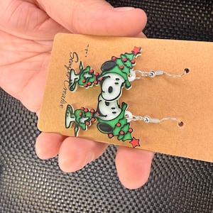 May include: A pair of Christmas-themed earrings featuring a cartoon character wearing a green Christmas tree hat. The earrings are white, black, and green with red star accents, and are displayed on a brown card.
