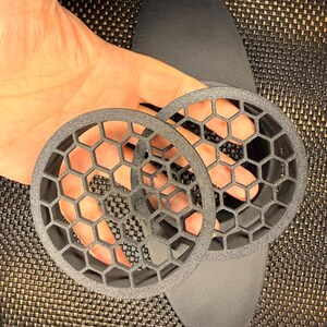 May include: Two circular black components featuring a raised honeycomb lattice pattern. These objects have a textured matte finish and are constructed with a rigid, open-grid structure. The design consists of a solid outer ring surrounding a hexagonal mesh.