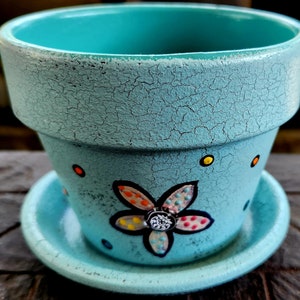 May include: A turquoise ceramic planter with a flower design and a matching saucer. The planter has a crackled finish and is decorated with colorful dots and a flower with a black outline.