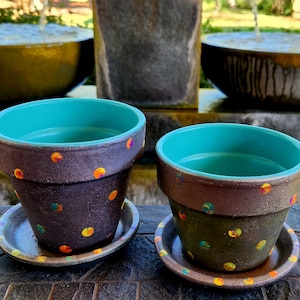 May include: Two terracotta flower pots with teal rims and a brown speckled base. The pots are decorated with colorful polka dots and have matching saucers.