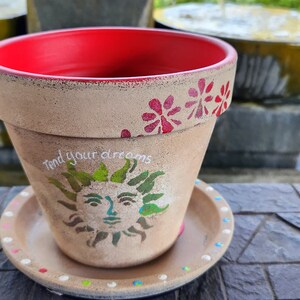 May include: A terracotta flower pot with a red rim and a painted sun design. The pot has the words "Find your dreams" written on it. The pot is sitting on a matching saucer.