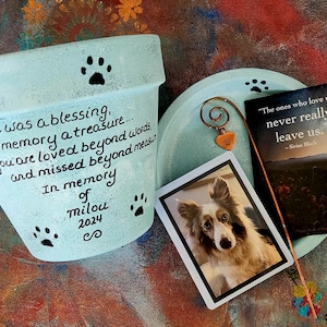May include: A light blue ceramic planter with a paw print design and a handwritten message that reads "your life was a blessing... your memory a treasure... you are loved beyond words and missed beyond measure. In memory of milou 2024 co". A small photo of a dog is attached to the planter with a metal heart charm.