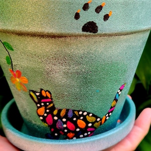 May include: A teal green terracotta planter with a black paw print, a black cat with colorful polka dots, and a single orange flower with green leaves painted on the side.