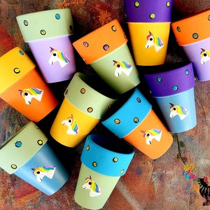 May include: A collection of colorful, hand-painted flower pots. Each pot features a unicorn design and a contrasting rim with polka dots. The pots are in shades of yellow, orange, green, purple, and blue, creating a whimsical and cheerful display.