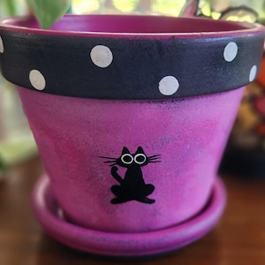 May include: A vibrant pink terracotta plant pot with a matching saucer. The rim is painted black with white polka dots. A black silhouette of a cat with large white eyes is centered on the front of the pot, which features a textured finish.