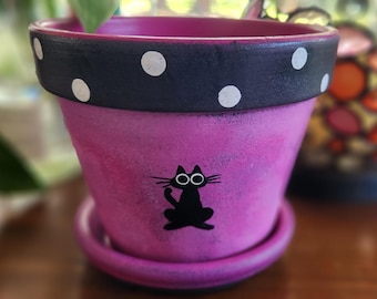 May include: A tapered terracotta plant pot with a matching saucer, featuring a vibrant magenta body and a black rim decorated with white polka dots. A small, stylized black cat silhouette with large circular eyes is painted on the front.
