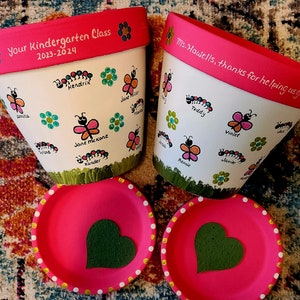 May include: Two white ceramic flower pots with pink rims, decorated with green, yellow, and blue paint. The pots have names and a caterpillar design. One pot says "Your Kindergarten Class 2023-2024" and the other says "Ms. Howells, thanks for helping us grow!" The pots are sitting on a pink and white patterned rug.