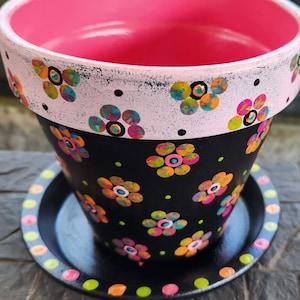 May include: Hand-painted flower pot with a black base and a pink interior. The pot features colorful floral designs and a white rim with more flowers. A matching saucer with a floral border completes the set. Perfect for adding a touch of color to any space.