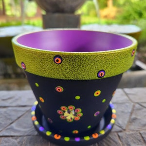 May include: A black and green Halloween-themed planter with a purple interior. The planter has a green rim with orange and black eyes painted on it. The main body of the planter is black with white polka dots and orange and black eyes. The planter has a matching saucer with a black rim and white polka dots.