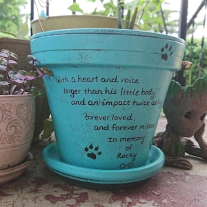 May include: A turquoise ceramic flower pot with a matching saucer, featuring black hand-painted paw prints and the text: With a heart and voice larger than his little body and an impact twice as big, forever loved, and forever missed, In memory of Rocky.
