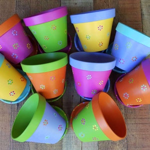May include: A collection of nine colorful terracotta flower pots with a white daisy pattern. The pots are painted in various shades of green, blue, orange, and pink.