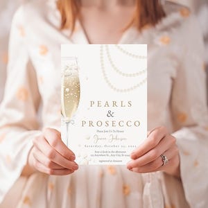 May include: A white invitation card with gold lettering that reads "Pearls & Prosecco" and features a glass of champagne with pearls. The invitation is for a party for Grace Johnson on Saturday, October 22nd.
