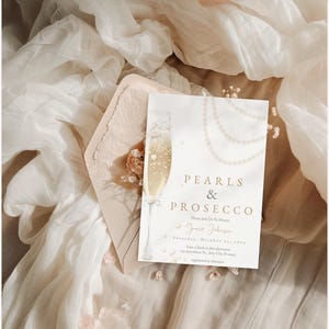 May include: A white invitation card with gold lettering that reads "Pearls & Prosecco". The card features a watercolor illustration of a champagne flute and a pearl necklace.