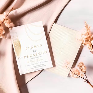 May include: A white invitation card with gold lettering that reads "Pearls & Prosecco". The card features a champagne flute with pearls and a floral design. The invitation is for a party on Saturday, October 29th.