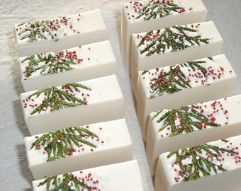 Pode incluir: Barras de sabão retangulares brancas decoradas com raminhos verdes e granulado vermelho. Os sabonetes estão dispostos em fileiras, mostrando um design festivo e natural. Estes sabonetes são provavelmente feitos à mão e adequados para presentear ou uso pessoal.