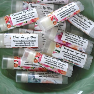 May include: A close-up of a bowl filled with various flavored lip glosses. The lip glosses are in clear tubes with colorful labels that list the flavors: Caramel, Chat Tea, Peppermint, Tangerine, Vanilla, and Strawberry. Each label includes the ingredients: almond oil, beeswax, shea butter, and essential oil or flavor oil. The labels also include the website address: www.joansgardens.etsy.com.