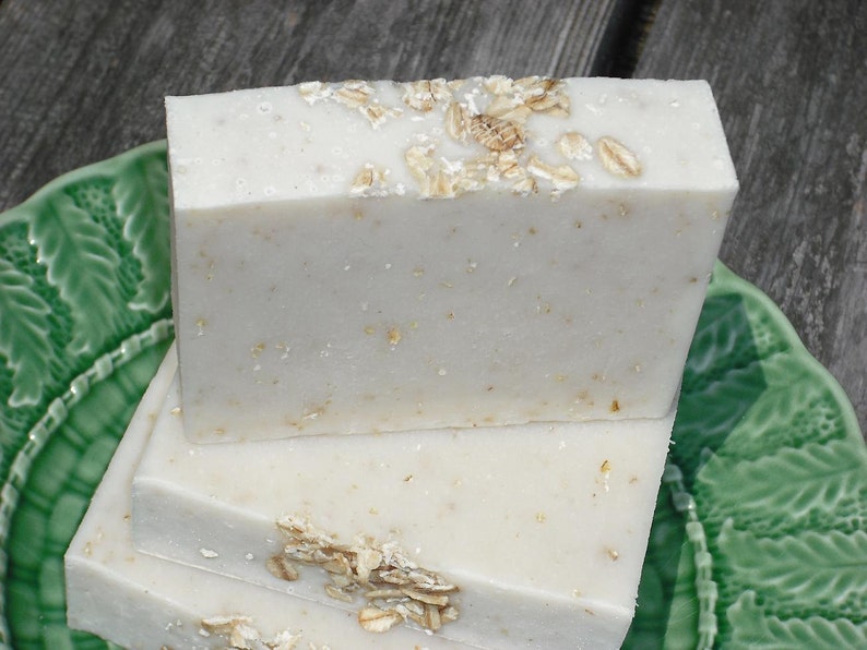 May include: Three bars of white soap with oatmeal flakes on a green plate. The soap is stacked on top of each other.