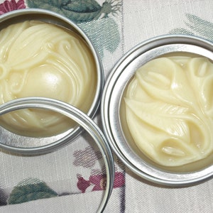 May include: Two silver tins with lids containing a white, creamy substance with a floral design. The substance is likely a hand cream or body butter.