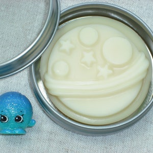 May include: A small tin container with a lid, containing a white, round, scented soap with a design of a crescent moon, stars, and a comet. The soap is in a tin with a silver lid.