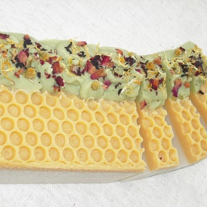 May include: A bar of handmade soap with a green layer on top and a yellow honeycomb pattern on the bottom. The green layer is decorated with dried flowers and herbs.