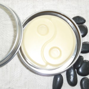May include: A round silver tin with a lid, containing a white, circular, solid soap with a yin yang symbol design. The tin is sitting on a white fabric surface with black stones scattered around it.