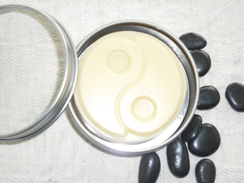 May include: A round silver tin with a lid, containing a white, circular, solid soap with a yin yang symbol design. The tin is sitting on a white fabric surface with black stones scattered around it.
