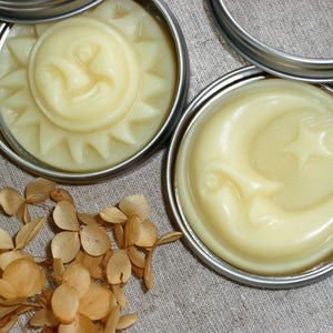 May include: Two silver tins with lids containing a yellow, creamy substance. The substance in the left tin is shaped like a sun with a smiling face. The substance in the right tin is shaped like a crescent moon with a star.