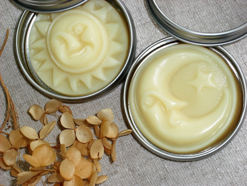 May include: Two silver tins with lids containing a yellow, creamy substance. The substance in the left tin is shaped like a sun with a smiling face. The substance in the right tin is shaped like a crescent moon with a star.