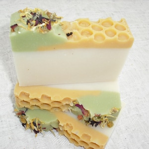 May include: Two bars of handmade soap, one with a honeycomb pattern and a green layer, the other with a white layer and a green layer with dried flowers and herbs.