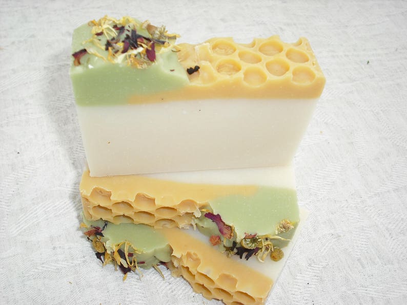 May include: Two bars of handmade soap, one with a honeycomb pattern and a green layer, the other with a white layer and a green layer with dried flowers and herbs.