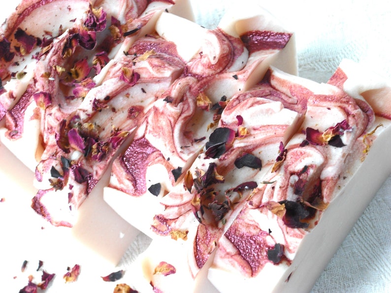 May include: Four bars of handmade soap with a swirl design in shades of white and pink. The soap is decorated with dried rose petals.