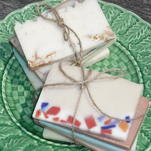 May include: A stack of handmade soap bars wrapped in twine and displayed on a green ceramic plate. The soap bars have a variety of colors and patterns, including white, brown, and red.