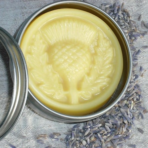 May include: A round tin container with a lid, filled with a yellow balm. The balm has a raised design of a thistle flower. The tin is sitting on a surface covered in dried lavender flowers.