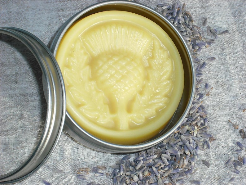 May include: A round tin container with a lid, filled with a yellow balm. The balm has a raised design of a thistle flower. The tin is sitting on a surface covered in dried lavender flowers.