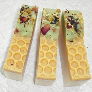 May include: Three bars of handmade soap with a green top layer and a yellow honeycomb pattern on the bottom layer. The green layer is decorated with dried flowers and herbs.