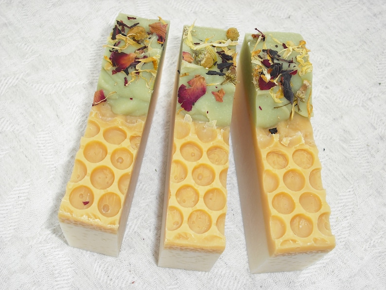 May include: Three bars of handmade soap with a green top layer and a yellow honeycomb pattern on the bottom layer. The green layer is decorated with dried flowers and herbs.