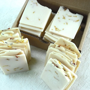 May include: A cardboard box filled with handmade soap bars. The soap bars are white with dried flowers and herbs embedded in them. The soap bars are stacked in two piles.