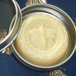 May include: A round tin container with a lid, containing a pale yellow balm with a bee design embossed on the surface.