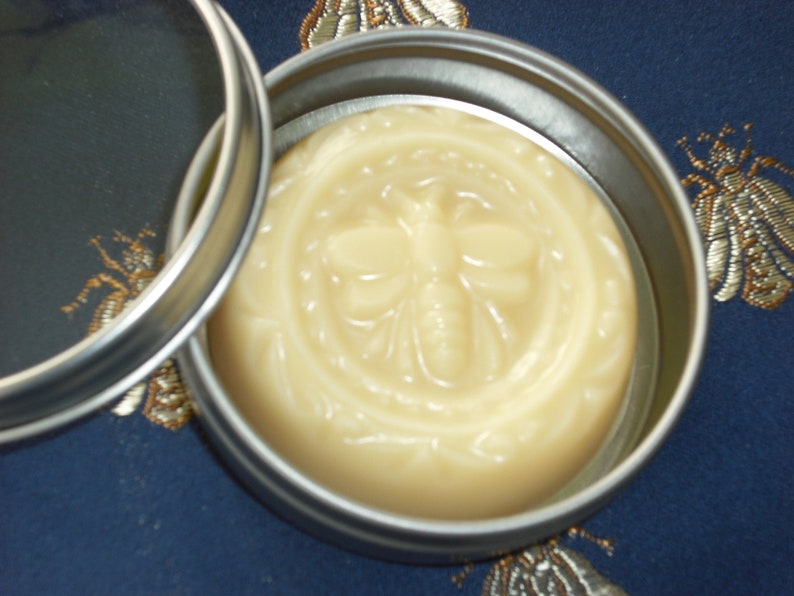 May include: A round tin container with a lid, containing a pale yellow balm with a bee design embossed on the surface.