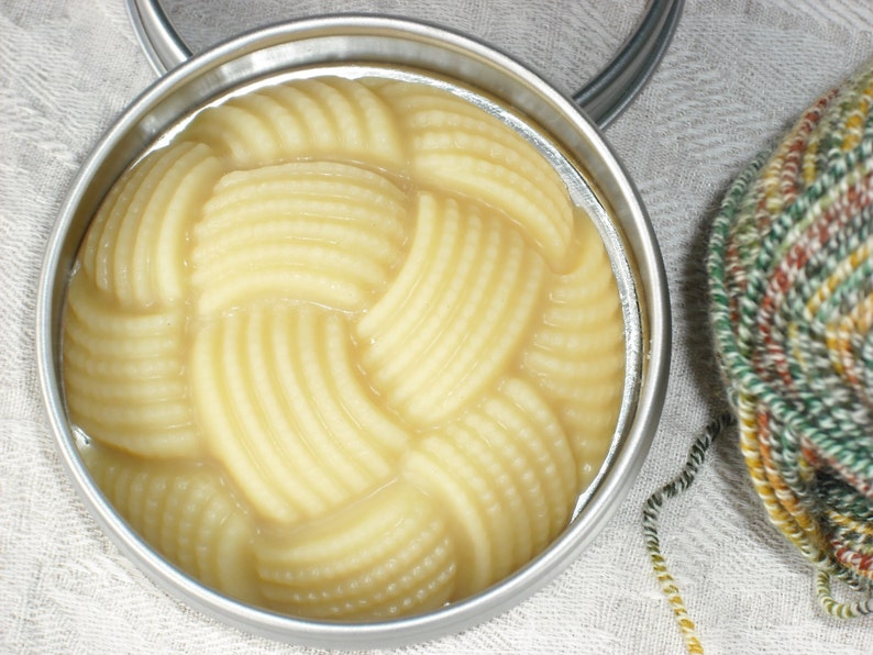 May include: A round metal tin filled with a thick, creamy, pale yellow substance. The substance is arranged in a woven pattern.