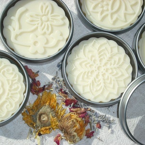 May include: Five round, white, decorative soap bars in metal tins. The soap bars have intricate floral designs pressed into them. The tins are arranged on a white fabric surface with dried flowers and petals scattered around them.