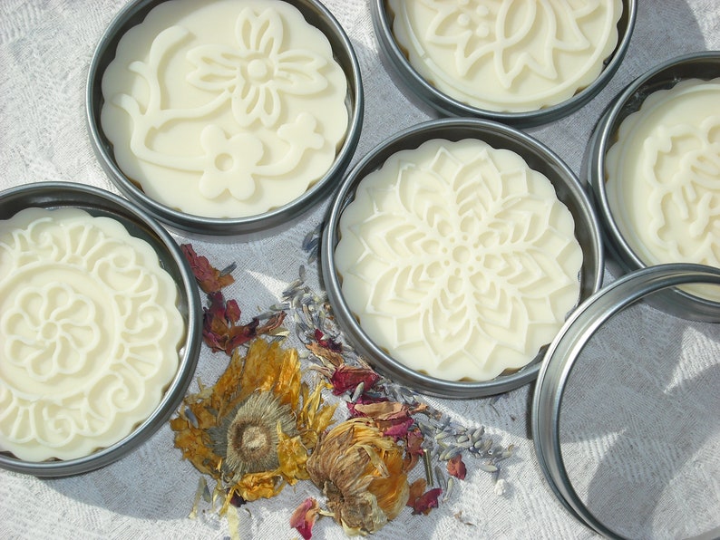 May include: Five round, white, decorative soap bars in metal tins. The soap bars have intricate floral designs pressed into them. The tins are arranged on a white fabric surface with dried flowers and petals scattered around them.