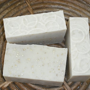 May include: Three rectangular bars of handmade soap. Two bars are wider, with a textured surface and embedded flecks. The third bar is narrower and taller, with a decorative circular pattern. The soaps are light beige.