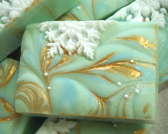 Pode incluir: Uma barra de sabão retangular num tom verde menta pálido, com um emblema de floco de neve branco moldado no topo. A superfície é decorada com detalhes metálicos dourados em espiral e pequenas contas esféricas brancas incrustadas.