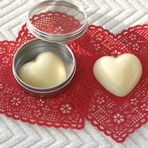 May include: Two heart-shaped containers of white body butter, one in a silver tin with a lid, and the other on a red lace doily.